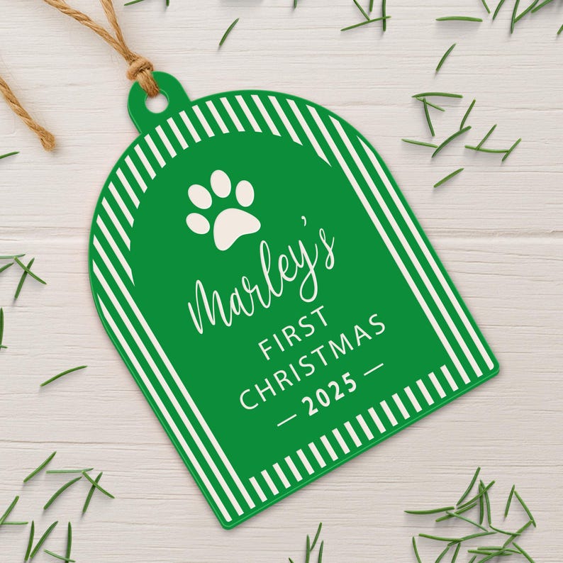 May include: A green Christmas ornament with white diagonal stripes and the text "Marley's First Christmas - 2025". The ornament has a paw print and is tied with a brown string.