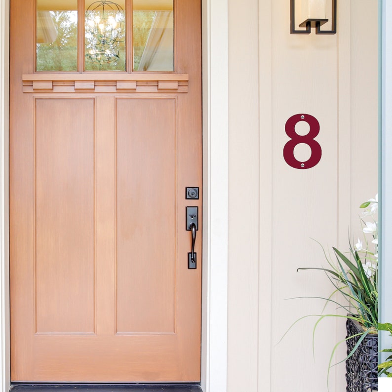 May include: A burgundy metal house number eight is fixed to a cream-coloured wall with two silver screws. A wooden door is to the left. The number is a stylish addition to any home.