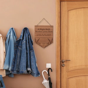 May include: A wooden sign with the text "A Night Shift Worker Lives Here" and "PLEASE DO NOT KNOCK OR RING THE DOORBELL". The sign is brown with a decorative floral design and a heart. It hangs on a wall near a door.