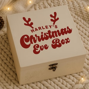 May include: A cream-colored wooden Christmas Eve box with red text that reads "Harley's Christmas Eve Box." The box has a decorative antler design and a metal clasp. The box is sitting on a cream-colored knitted blanket.