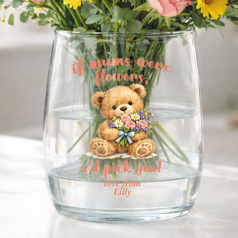 May include: A clear glass vase with a teddy bear illustration holding flowers. The text on the vase reads "if mums were flowers, I'd pick you!" and "love from Lilly". The vase contains water and a bouquet of flowers.