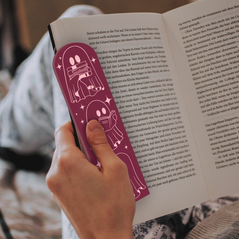 May include: A burgundy bookmark featuring a pink outline of three ghosts reading books. The ghosts are holding books and have stars around them. The bookmark is being used to mark a page in a book.
