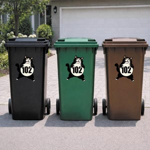 May include: Three wheeled trash bins in black, green, and brown. Each bin has a sticker of a black and white cat holding a white circle with the number 102. The bins are on a paved surface.