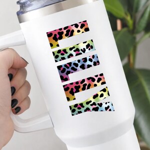 May include: White travel mug with a handle and a silver lid. The word "PINK" is displayed vertically in colorful leopard print letters. The leopard print includes black spots and a rainbow of colors.