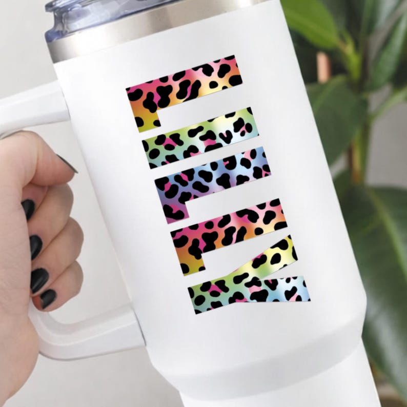 May include: White travel mug with a handle and a silver lid. The word "PINK" is displayed vertically in colorful leopard print letters. The leopard print includes black spots and a rainbow of colors.