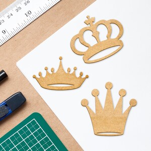 May include: Three wooden crown cutouts in various designs. One crown has a cross on top, another has a series of small points, and the third has five rounded points. The crowns are light brown and sit on a white surface.