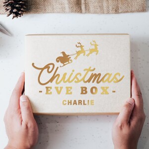 May include: A beige box with a gold foil design featuring Santa Claus in a sleigh pulled by reindeer. The text "Christmas Eve Box - CHARLIE" is printed on the box.