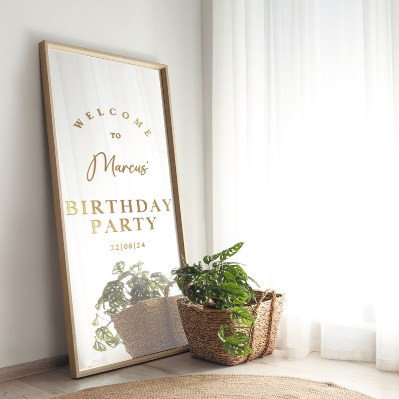 May include: A gold mirror with a wooden frame, reflecting a green plant in a wicker basket. The mirror has the text "Welcome to Marcus' Birthday Party 22|08|24" on it.