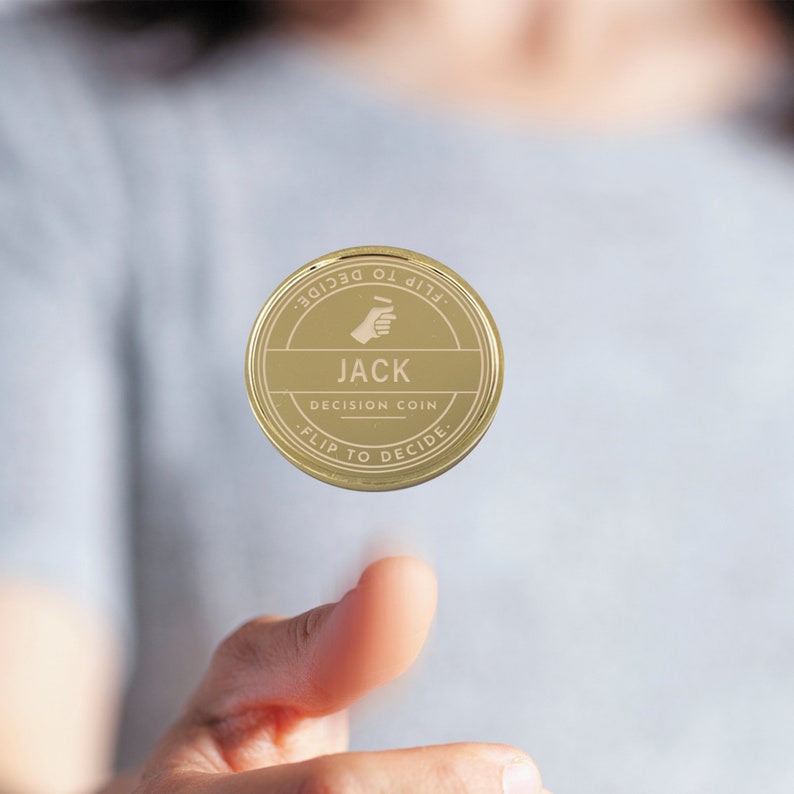 Puede incluir: Una moneda dorada de decisi&oacute;n con el nombre "JACK" en el centro. La moneda tiene las palabras "FLIP TO DECIDE" alrededor del borde y un gr&aacute;fico de mano. La moneda est&aacute; en el aire, sostenida por un dedo.