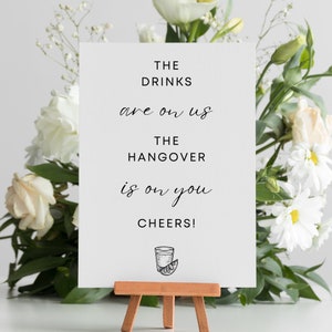 May include: White sign with black text that reads "The drinks are on us. The hangover is on you. Cheers!" with a black and white illustration of a shot glass with a lime wedge below the text.