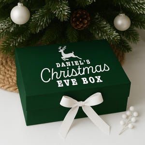 Pu&ograve; includere: Una scatola verde scuro della vigilia di Natale con un fiocco bianco e la scritta "Daniel's Christmas Eve Box" in bianco. La scatola presenta una grafica di renna sopra il testo. La scatola &egrave; decorata con un fiocco bianco ed &egrave; posta davanti a un albero di Natale.