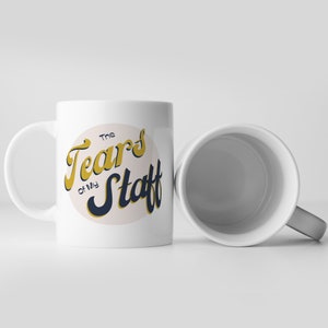 May include: White ceramic mug with a black and gold design that says "The Tears of My Staff".