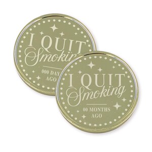 May include: Two round, olive-green coins with gold rims. The text "I QUIT Smoking" is centered on each coin. One coin reads "000 DAY AGO" and the other reads "00 MONTHS AGO". Each coin has a dotted border and star accents.