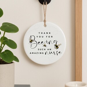 May include: A round, white ceramic ornament with the text "THANK YOU FOR Bee-ing SUCH AN AMAZING nurse." The design includes illustrations of bees. The ornament hangs from a natural twine string.