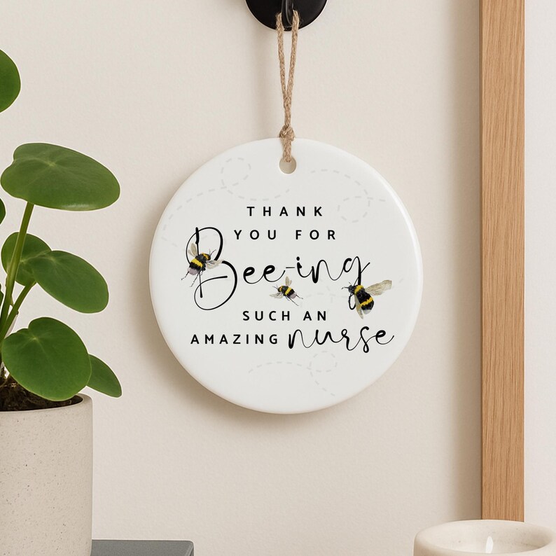 May include: A round, white ceramic ornament with the text "THANK YOU FOR Bee-ing SUCH AN AMAZING nurse." The design includes illustrations of bees. The ornament hangs from a natural twine string.