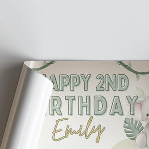 Puede incluir: Un p&oacute;ster de cumplea&ntilde;os con el texto "HAPPY 2ND BIRTHDAY Emily" en letras verdes y doradas. El dise&ntilde;o incluye un elefante de dibujos animados y follaje verde. El p&oacute;ster est&aacute; parcialmente enrollado, revelando un borde blanco.