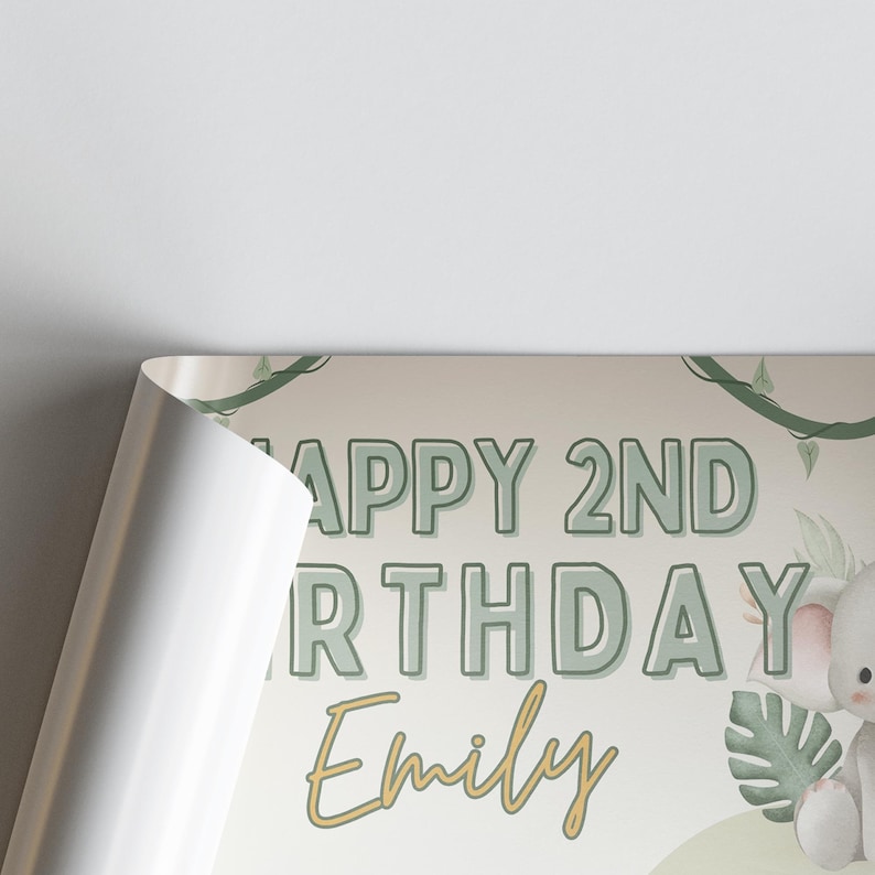 Puede incluir: Un p&oacute;ster de cumplea&ntilde;os con el texto "HAPPY 2ND BIRTHDAY Emily" en letras verdes y doradas. El dise&ntilde;o incluye un elefante de dibujos animados y follaje verde. El p&oacute;ster est&aacute; parcialmente enrollado, revelando un borde blanco.