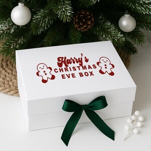 May include: A white Christmas Eve box tied with a dark green bow. The box features the text "Harry's CHRISTMAS EVE BOX" in red, and two gingerbread man graphics. The box is decorated with Christmas ornaments and a pine cone.