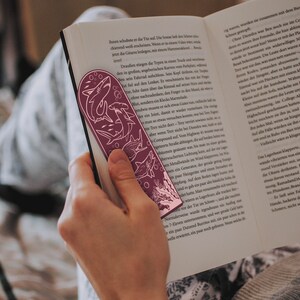 May include: A pink bookmark with a white outline of various sea creatures, including sharks, a hammerhead shark, and coral. The bookmark is being used to mark a page in a book.