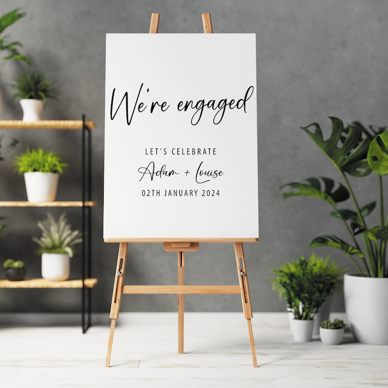 May include: White easel sign with black text announcing an engagement. The text reads "We're engaged" followed by "Let's celebrate Adam + Louise 02th January 2024".