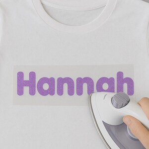 May include: A white t-shirt with a purple glitter iron-on design that says "Hannah" being ironed with a white iron.