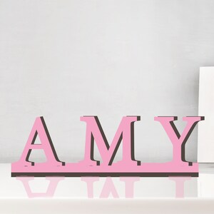 May include: Pink wooden letters spelling out the name "AMY" with black outlines. The letters are standing on a white surface.