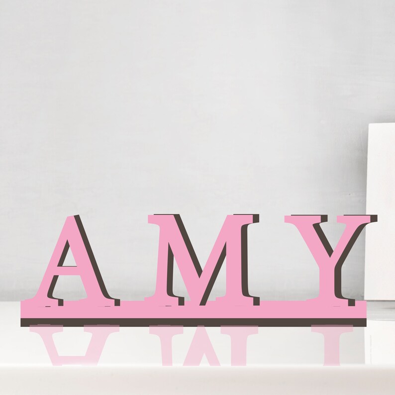 May include: Pink wooden letters spelling out the name "AMY" with black outlines. The letters are standing on a white surface.