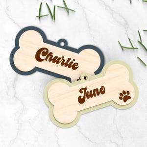 Pode incluir: Duas etiquetas de madeira em forma de osso de c&atilde;o. Uma &eacute; azul marinho com "Charlie" em escrita castanha. A outra &eacute; verde claro com "Juno" e uma pegada castanha. Ambas t&ecirc;m um centro com gr&atilde;o de madeira natural.
