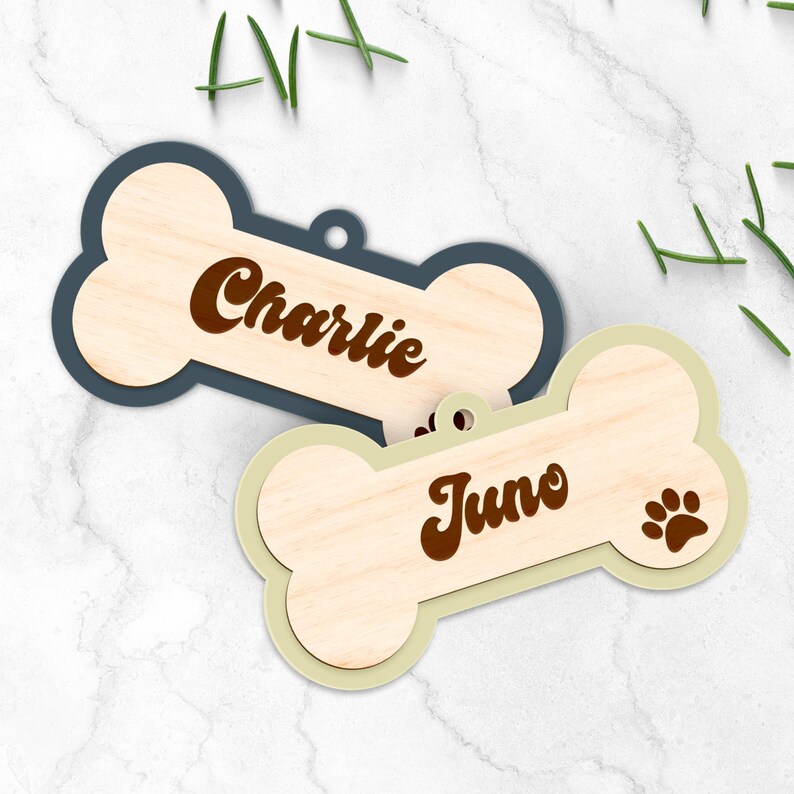 Pode incluir: Duas etiquetas de madeira em forma de osso de c&atilde;o. Uma &eacute; azul marinho com "Charlie" em escrita castanha. A outra &eacute; verde claro com "Juno" e uma pegada castanha. Ambas t&ecirc;m um centro com gr&atilde;o de madeira natural.