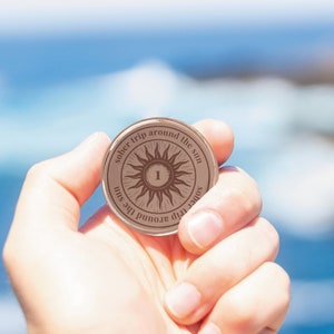 Op de afbeelding: Een ronde, bronskleurige soberheidsmunt die in een hand wordt gehouden. De munt heeft een zonnesterontwerp met het nummer "1" in het midden en de woorden "sober trip around the sun" rond de rand. De achtergrond is een vage weergave van de oceaan.