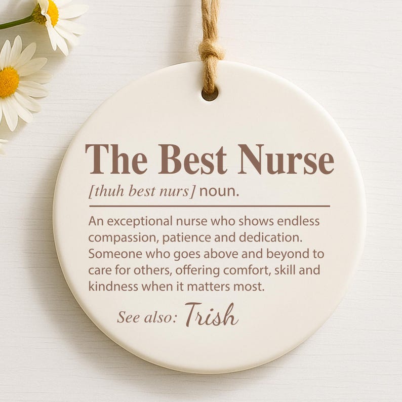 May include: A round, off-white ceramic ornament with the text "The Best Nurse" in brown. The text includes a definition of a nurse and the name "Trish". A natural twine loop is at the top for hanging.