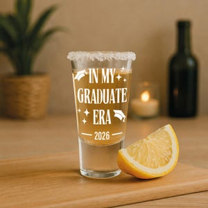 May include: A clear shot glass with a salted rim, filled with a golden liquid. The text "IN MY GRADUATE ERA 2026" is printed in white. A lemon wedge sits next to the glass on a wooden surface.