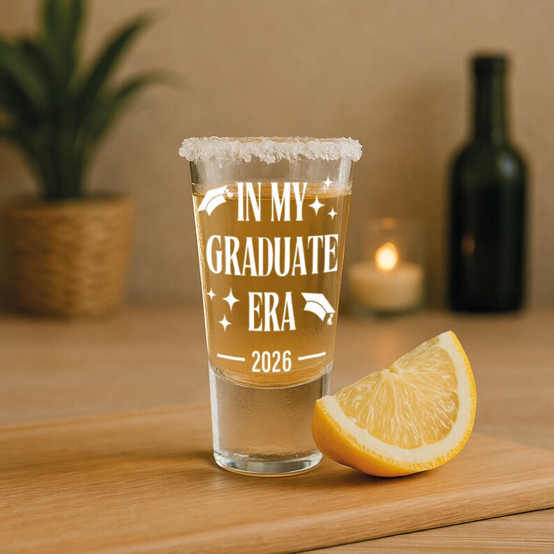 May include: A clear shot glass with a salted rim, filled with a golden liquid. The text "IN MY GRADUATE ERA 2026" is printed in white. A lemon wedge sits next to the glass on a wooden surface.