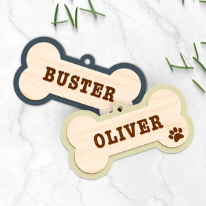 Peut inclure: Deux &eacute;tiquettes en bois en forme d'os de chien. L'une est bleu marine avec le nom "BUSTER" en marron. L'autre est vert clair avec le nom "OLIVER" et une empreinte de patte en marron. Les deux ont un trou pour l'attache au collier.