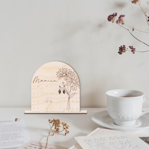 May include: Wooden earring holder with a floral design and the name "Monica" engraved on it. The holder has multiple holes for earrings and is shaped like an arch.
