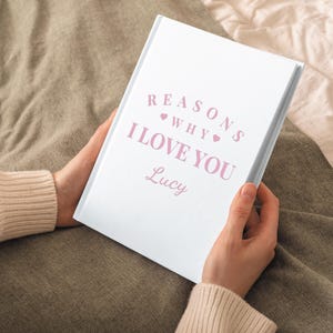 Może przedstawiać: Biały dziennik z tekstem "Reasons Why I Love You" w r&oacute;żowych literach z dwoma małymi r&oacute;żowymi sercami. Imię "Lucy" jest napisane r&oacute;żowym kursywą pod tekstem.