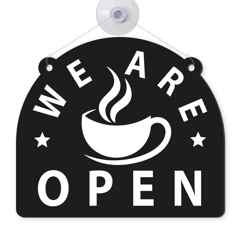 May include: Black and white cafe sign with the words "WE ARE OPEN" in a semi-circular arrangement. A white coffee cup with steam is in the center. Two white stars flank the cup. The sign is attached to a clear suction cup.