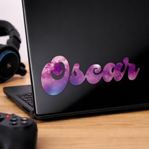 Może przedstawiać: Naklejka z napisem "Oscar" w kursywie, z fioletowo-niebieskim wzorem galaktyki, jest naklejona na czarnym laptopie. Naklejka ma wirujący efekt mgławicy z białymi gwiazdami.