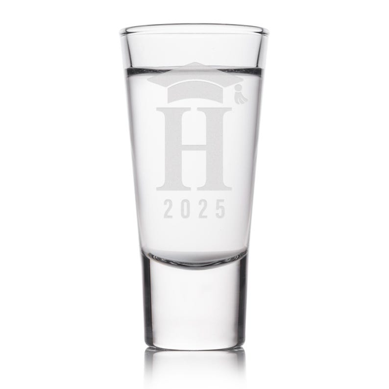 K&ouml;nnte beinhalten: Klares Schnapsglas mit einem Abschlusskappen-Design, dem Buchstaben "H" und der Jahreszahl "2025" auf der Vorderseite. Das Glas ist mit einer klaren Fl&uuml;ssigkeit gef&uuml;llt, vor wei&szlig;em Hintergrund.
