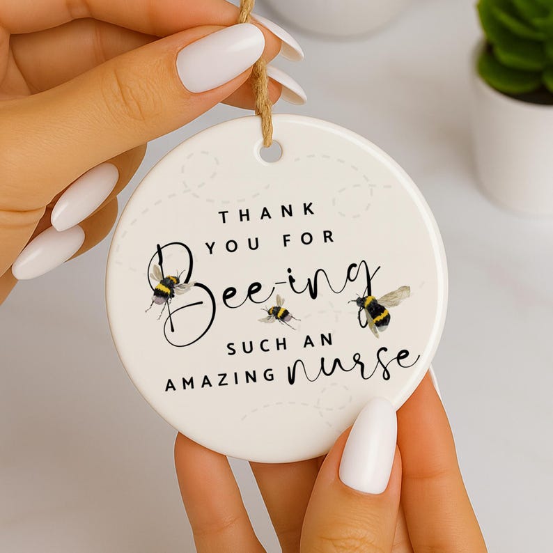 May include: A white ceramic ornament with the text "Thank You For Bee-ing Such An Amazing Nurse" and bee illustrations. The ornament is round with a hole at the top for hanging with a twine string.