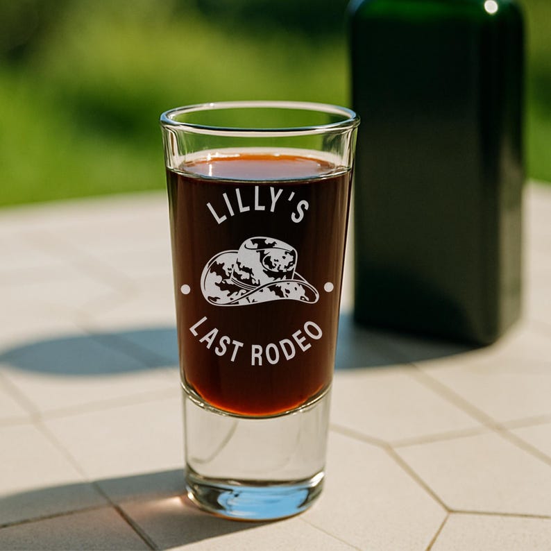 K&ouml;nnte beinhalten: Ein klares Schnapsglas, gef&uuml;llt mit einer dunklen Fl&uuml;ssigkeit, mit dem wei&szlig;en Text "Lilly's Last Rodeo". Eine Cowboyhut-Grafik befindet sich zwischen dem Text. Eine gr&uuml;ne Flasche ist im Hintergrund unscharf.