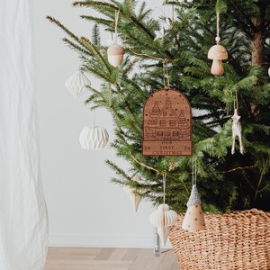 May include: A wooden Christmas ornament shaped like a house with a snowy roof and the text "Our First Christmas" on the bottom. The ornament is hanging on a Christmas tree with other ornaments.