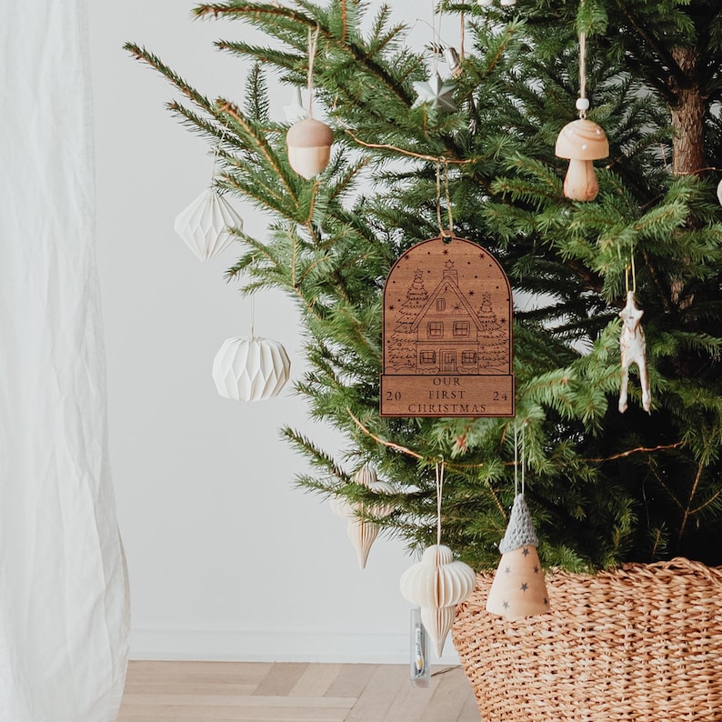 May include: A wooden Christmas ornament shaped like a house with a snowy roof and the text "Our First Christmas" on the bottom. The ornament is hanging on a Christmas tree with other ornaments.