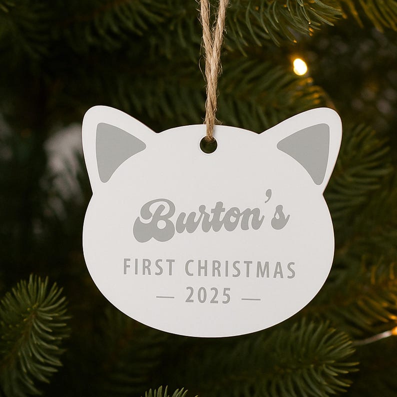 May include: A white cat-shaped Christmas ornament with gray accents. The ornament reads "Burton's FIRST CHRISTMAS - 2025 -" in gray lettering. It hangs from a natural twine string, set against a blurred green background.