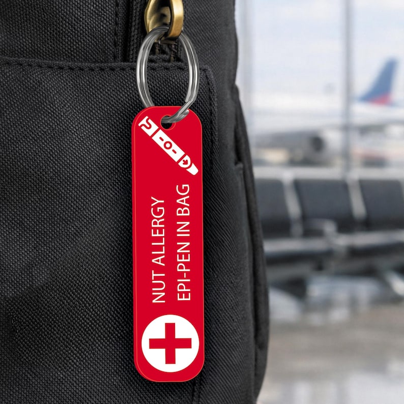 Op de afbeelding: Een rode rechthoekige sleutelhanger met de tekst "NUT ALLERGY EPI-PEN IN BAG" en een wit kruissymbool. De sleutelhanger is bevestigd aan een zwarte tas met een zilveren sleutelring. Bovenop staat een witte afbeelding van een EpiPen.