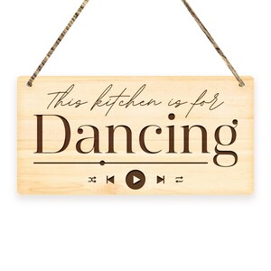 May include: Wooden sign with the text "This kitchen is for Dancing" in a script font. Below the text is a music player graphic with play, pause, skip, and shuffle icons. The sign is light brown with a natural wood grain and a twine hanger.
