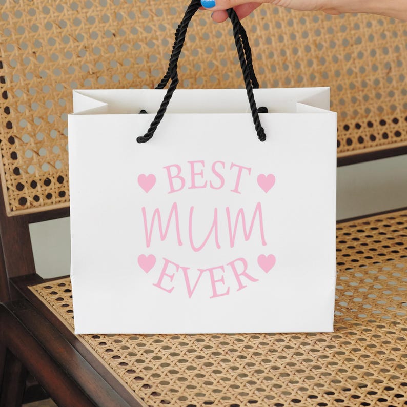 Op de afbeelding: Een witte cadeauzak met zwarte handvatten en roze tekst die "BEST MUM EVER" zegt met roze hartjes aan weerszijden van de tekst.