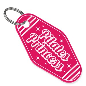 May include: A pink keychain with the words "Pilates Princess" in white script. The keychain has a retro motel style design with white star and stripe accents and a silver key ring.