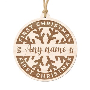 May include: A round wooden Christmas ornament with the words "FIRST CHRISTMAS" in a circle around a snowflake design. The center has the words "Any name" and the year "2025". A natural twine loop is attached for hanging.
