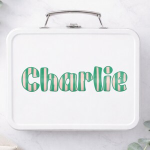 Op de afbeelding: Witte metalen lunchbox met een zilveren handvat. De naam "Charlie" is in grote, retro-stijl letters op de voorkant gedrukt. De letters zijn groen met roze verticale strepen, wat een speels en gepersonaliseerd ontwerp cre&euml;ert.
